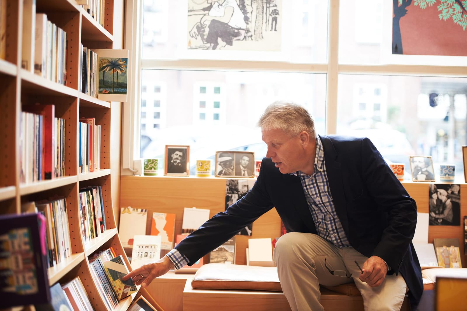 Homem selecionando um livro em uma prateleira de uma livraria, cercado por publicações e cartões ilustrados