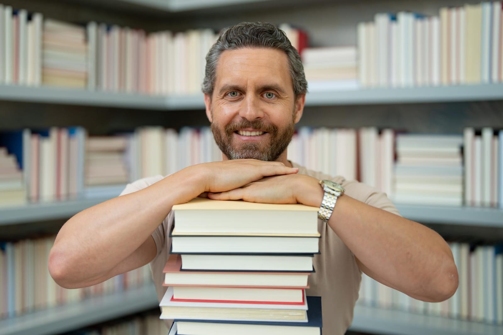 Homem sorridente apoiado em uma pilha de livros, diante de estantes cheias de livros em uma biblioteca