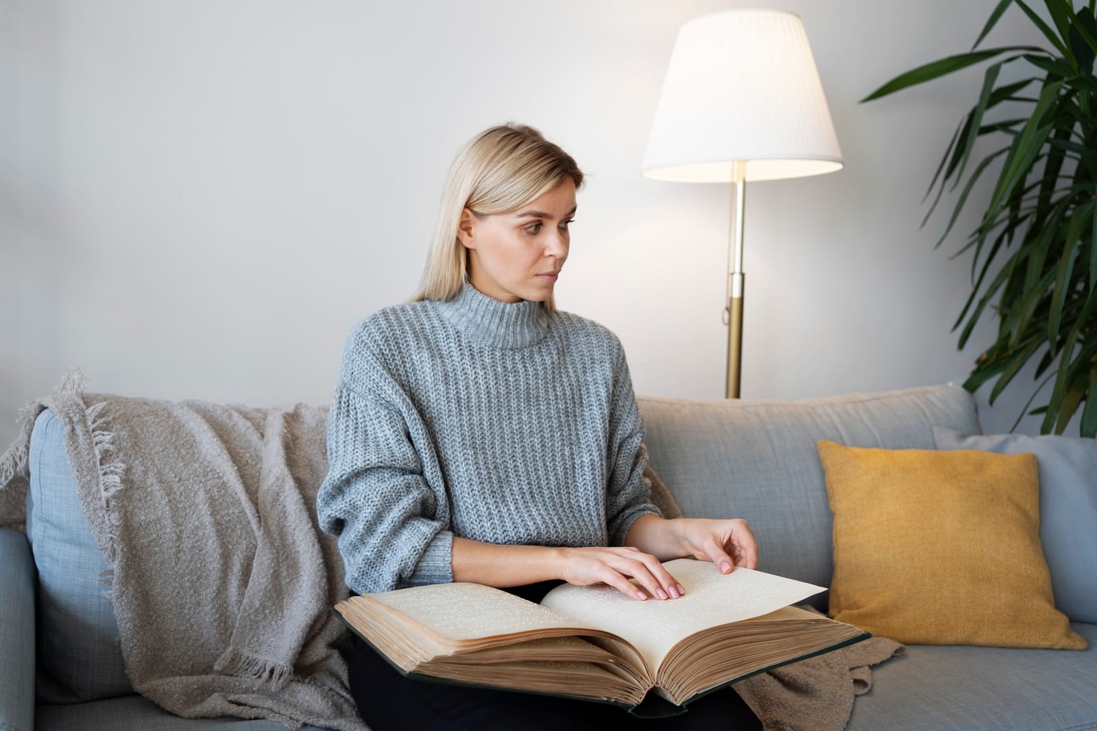 Mulher sentada no sofá de casa, lendo um livro grosso apoiado no colo, perto de uma luminária